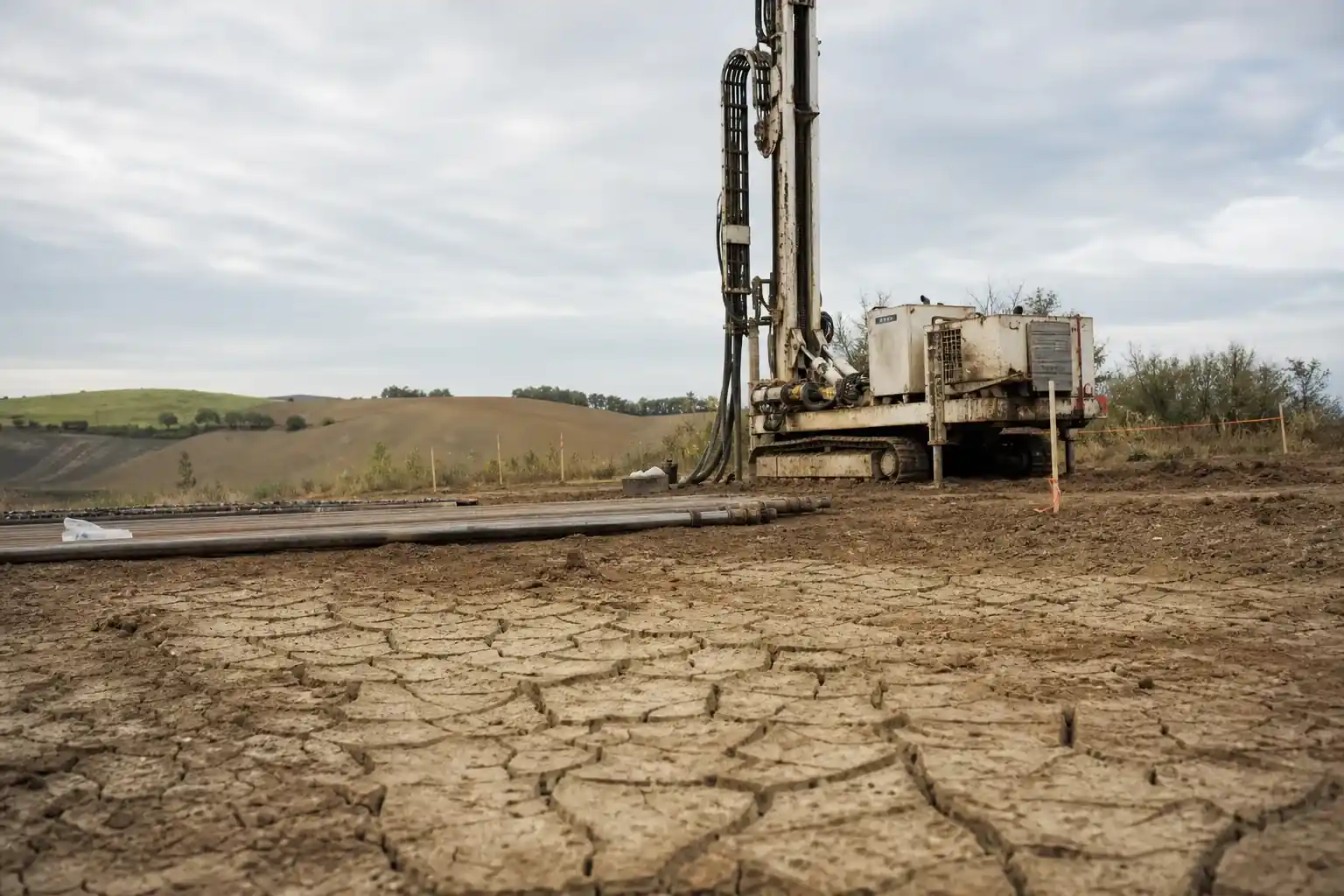Sondage géotechnique sur un terrain nu, foreuse compacte et tiges de forage posées au sol, argile fissurée par la sécheresse au premier plan